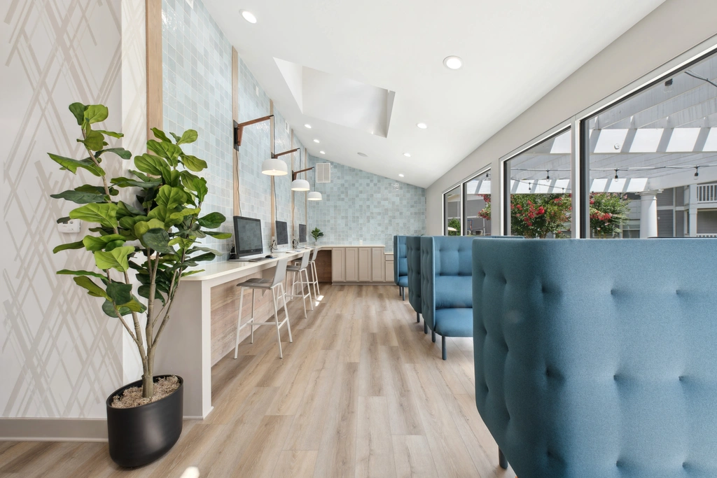 Bright resident lounge and workspace with booth seating, a row of computers, pendant lighting, large windows, and modern finishes.