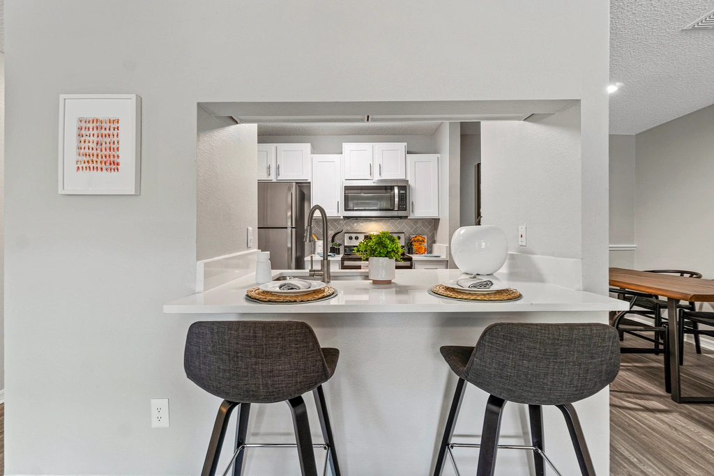 Modern kitchen with breakfast bar seating, featuring white cabinetry, stainless steel appliances, and a clean, open pass-through design into the living area.