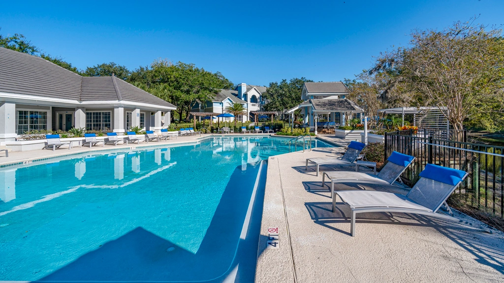 Resort-style swimming pool with surrounding lounge chairs, landscaped grounds, and adjacent clubhouse under a clear blue sky.