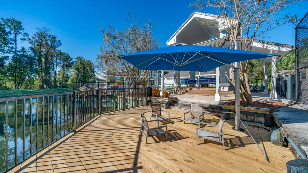 Outdoor lounge area with seating on a wooden deck, featuring shaded umbrellas, waterfront views, and access to a nearby clubhouse and amenities.