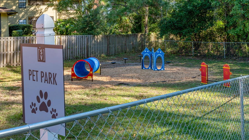 Fenced pet park with agility equipment, including tunnels and play structures, set within a landscaped outdoor area.