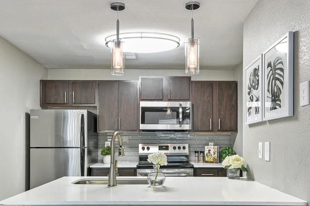 Modern apartment kitchen featuring dark wood cabinetry, stainless steel appliances, quartz countertops, a large kitchen island with undermount sink, glass tile backsplash, and contemporary pendant lighting.