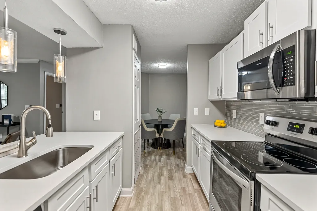 Bright modern apartment kitchen with white shaker cabinetry, stainless steel appliances, quartz countertops, gray tile backsplash, wood-style flooring, and pendant lighting overlooking a cozy dining area.