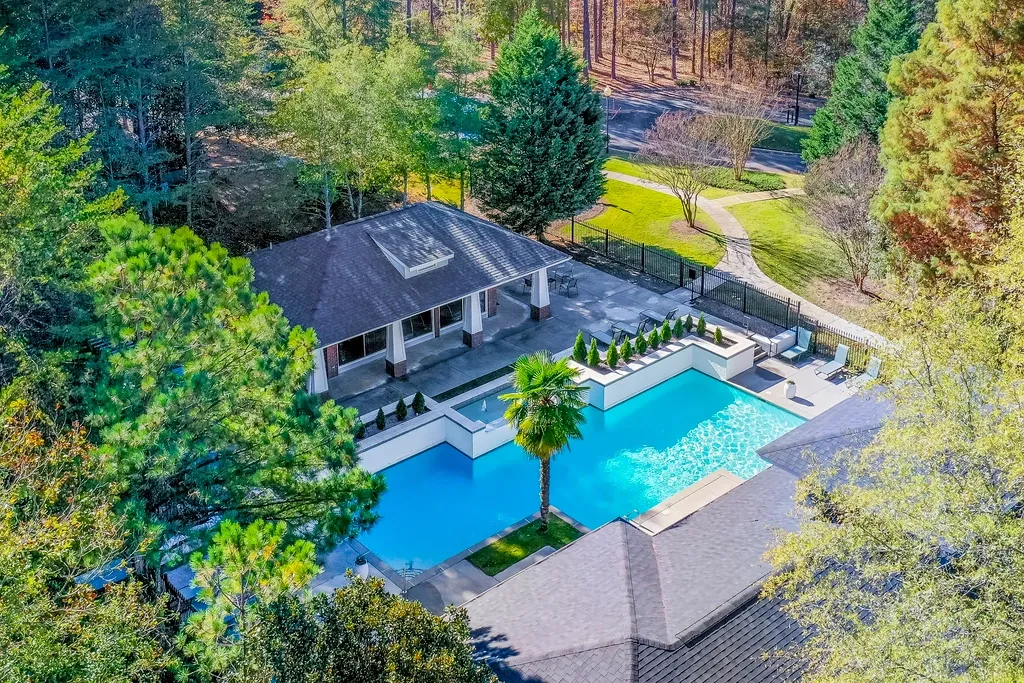 Aerial view of a resort-style apartment swimming pool surrounded by lush trees, landscaped walking paths, lounge seating, and a covered outdoor pavilion in a peaceful wooded setting.