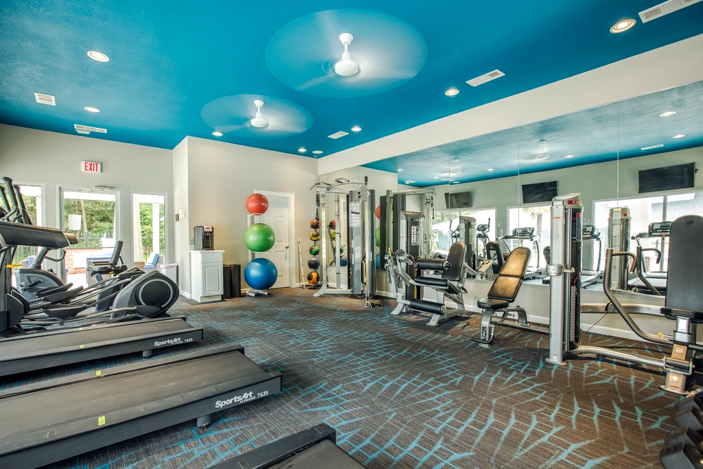 Modern fitness center featuring cardio and strength-training equipment, mirrored walls, free weights, exercise balls, wall-mounted TVs, and a vibrant blue ceiling with recessed lighting and ceiling fans.
