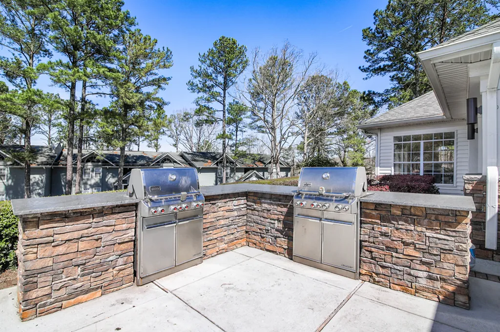 Chic outdoor BBQ area with stainless steel grills set against a rustic stone wall, surrounded by lush greenery and ample patio space.
