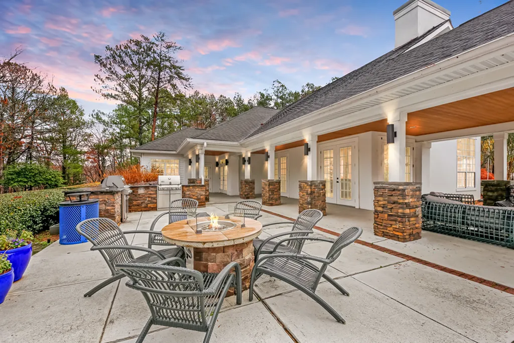 Elegant outdoor patio with cozy fire pit, stylish seating, and a modern BBQ grill, set against a charming house with stone columns and lush greenery.
