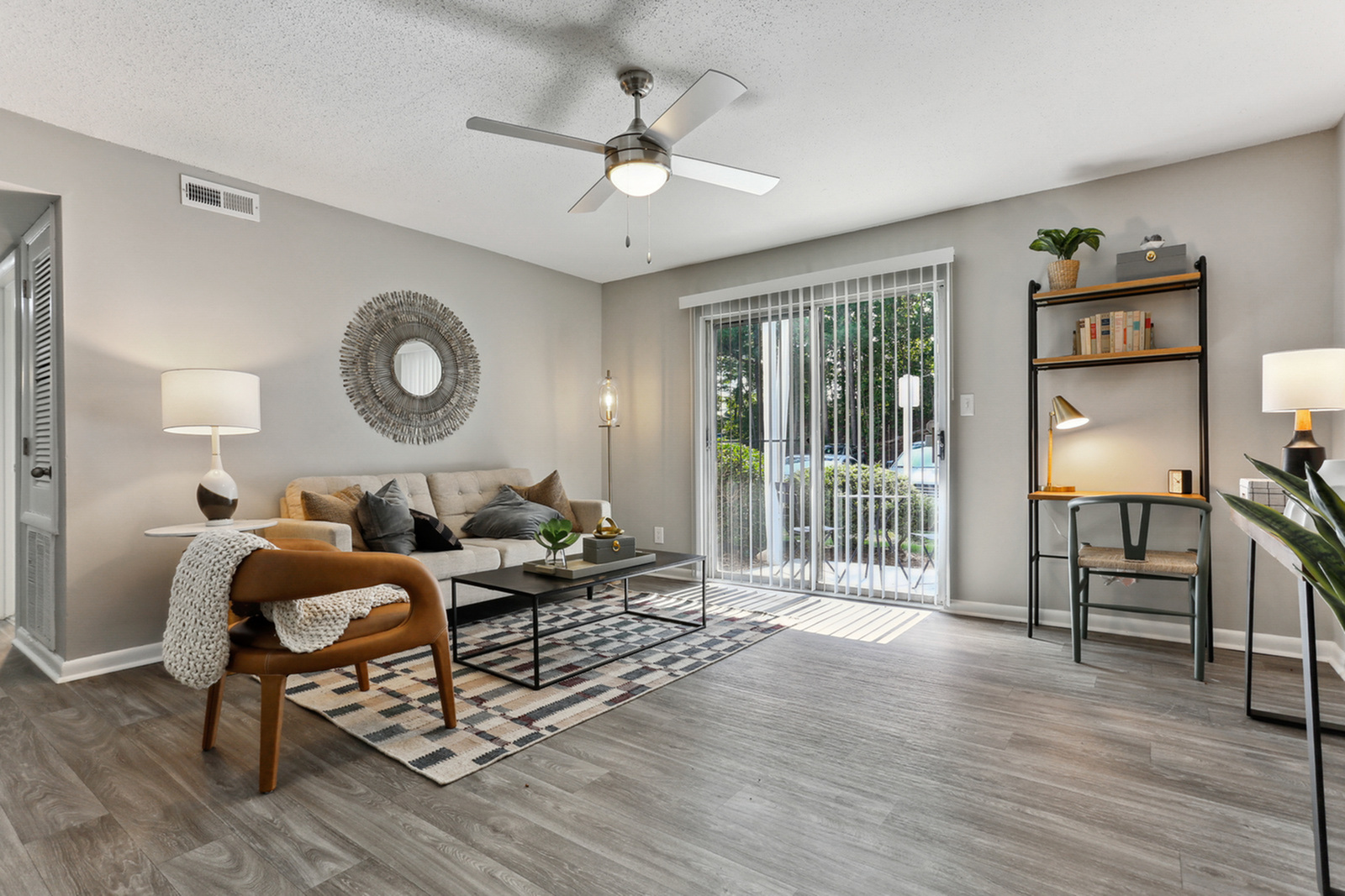 Elegant living room featuring a cozy sofa, chic round decorative mirror, modern ceiling fan, and abundant natural light from large sliding doors leading to a serene outdoor view.