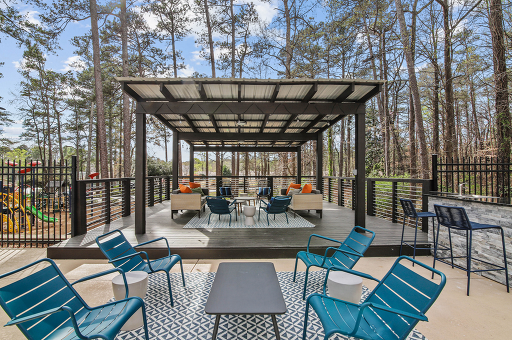 Charming outdoor seating area with stylish chairs and cozy sofas under a covered pergola, surrounded by lush trees and vibrant playground nearby.