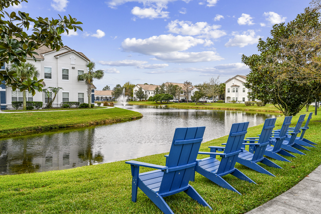 Captivating apartment lake view with blue Adirondack chairs, lush greenery, and elegant architecture under a sunny sky.