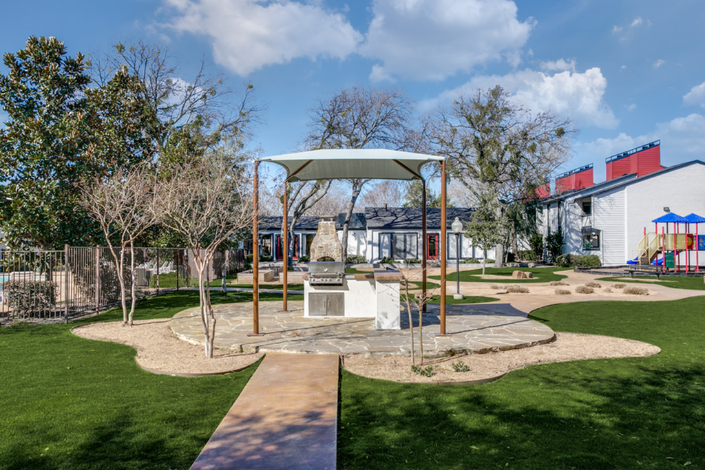 Inviting apartment community courtyard featuring a modern barbecue area, lush greenery, and a vibrant children\'s playground under a bright blue sky.