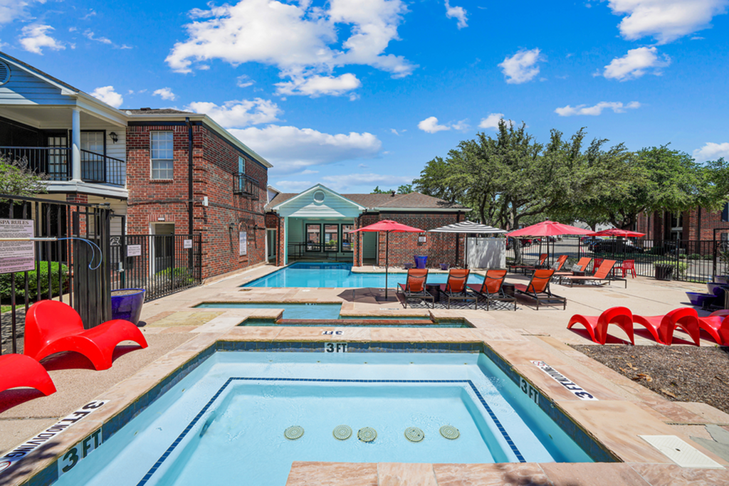 Inviting apartment pool area with elegant brick buildings, vibrant red loungers, and lush greenery under a bright blue sky.