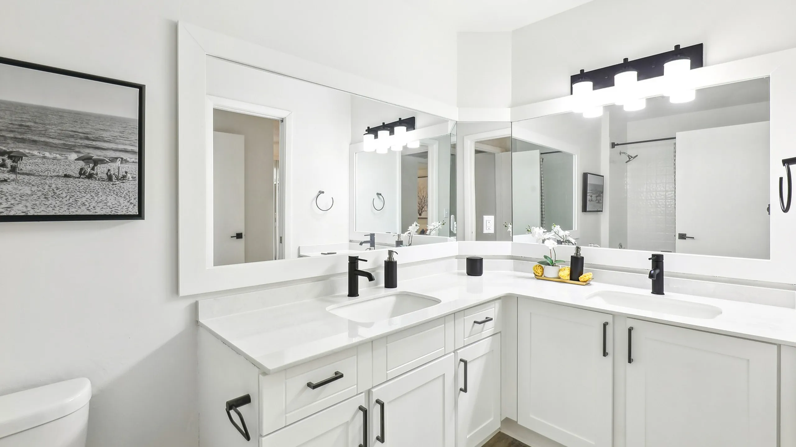 Modern white bathroom with corner vanity, black hardware, large mirrors, and bright lighting. Clean and minimalistic.