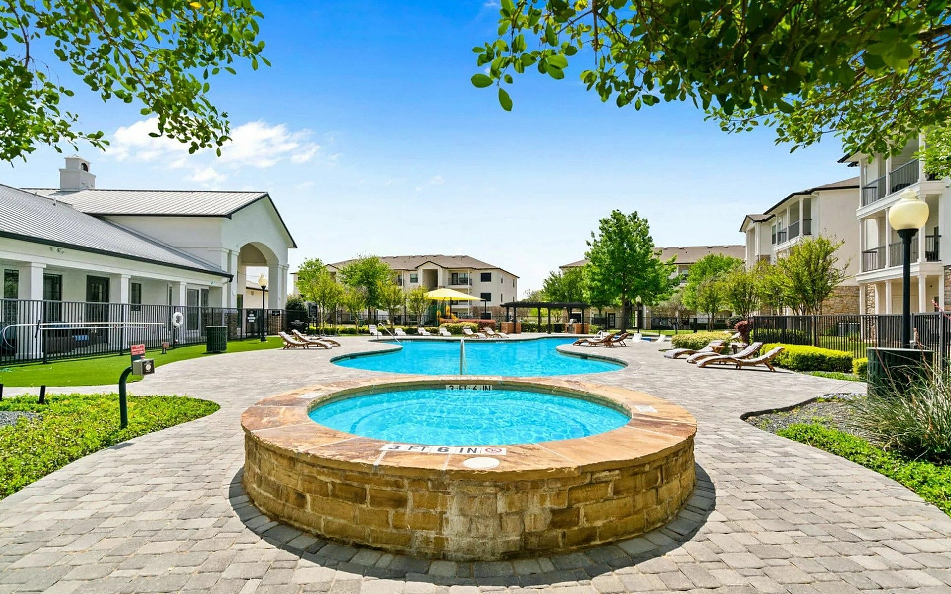 Inviting apartment complex with a luxurious outdoor pool area, lush greenery, and modern lounging chairs under a clear blue sky.