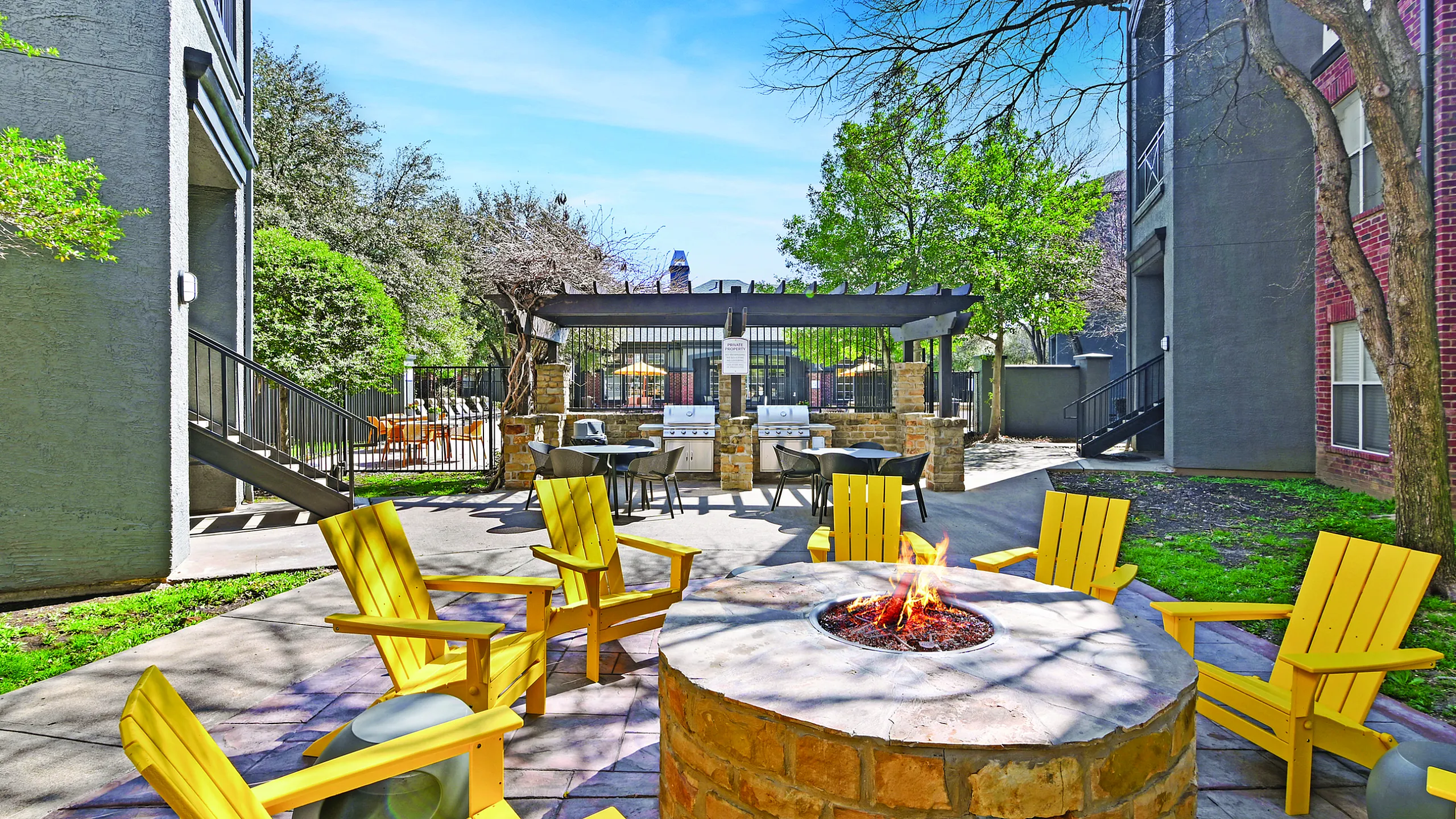 Charming outdoor patio with cozy yellow chairs surrounding a warm stone fire pit, complemented by a modern barbeque station and lush greenery.