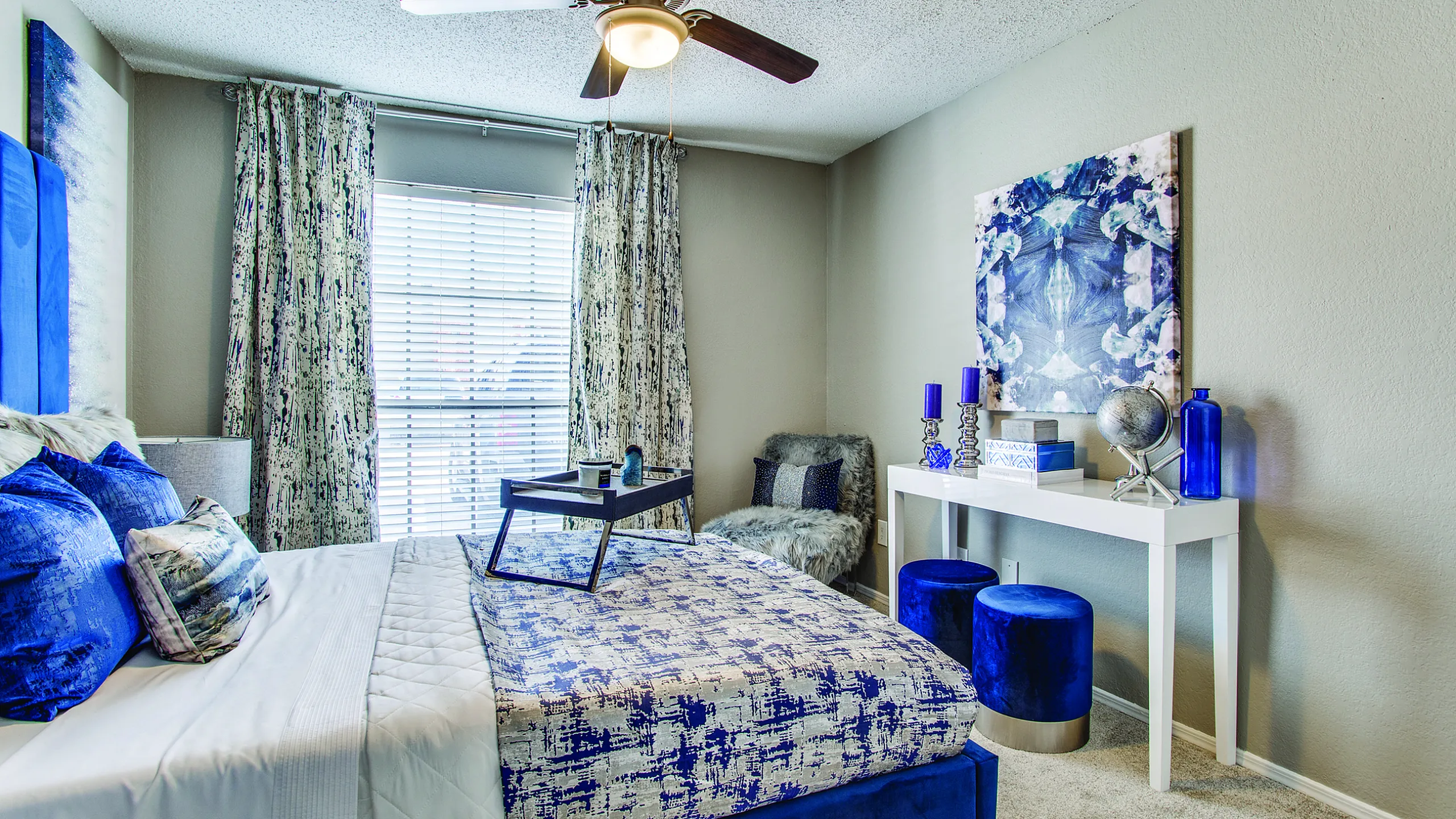 Elegant bedroom with a modern design, featuring a luxurious bed with blue accents, chic artwork, and a stylish ceiling fan.