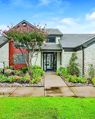Charming residential building with lush landscaping, vibrant flowers, and a welcoming entrance under a clear blue sky.