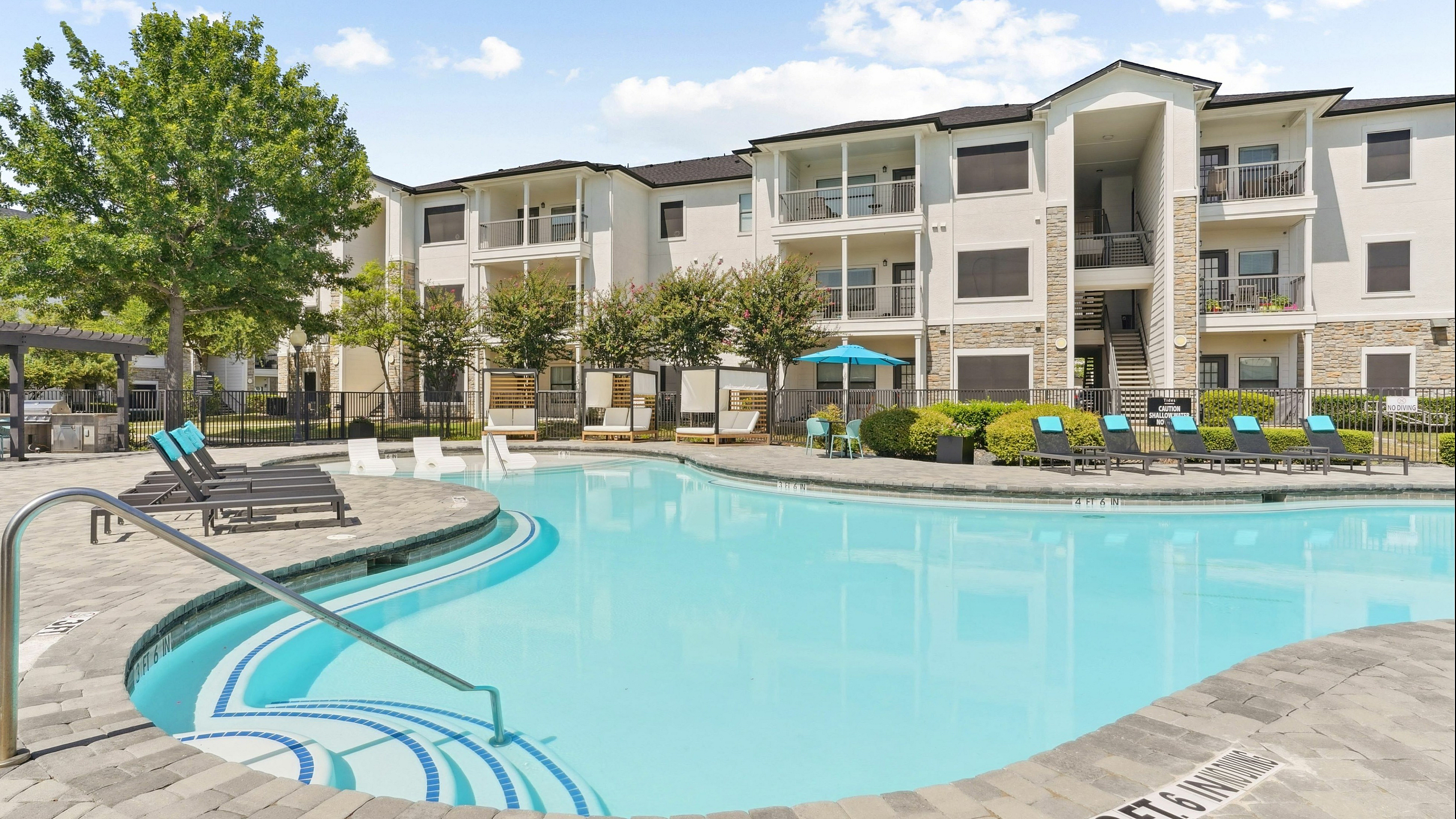 Luxurious apartment complex with a sparkling pool, stylish lounging area, and modern outdoor seating under a clear blue sky.