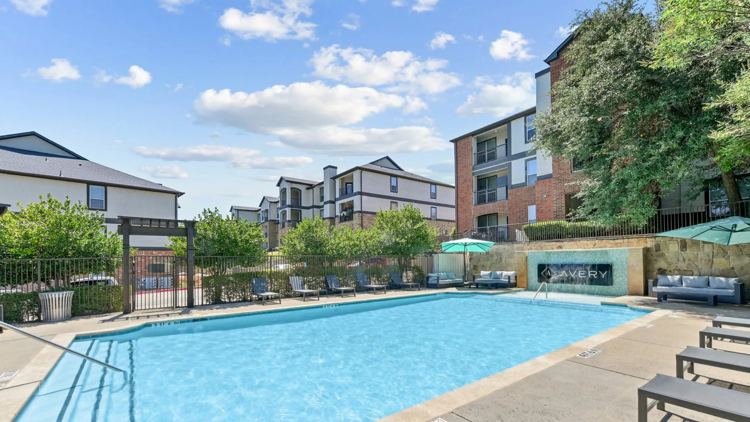Charming apartment complex with a sparkling pool and cozy seating area, surrounded by lush greenery under a sunny sky.