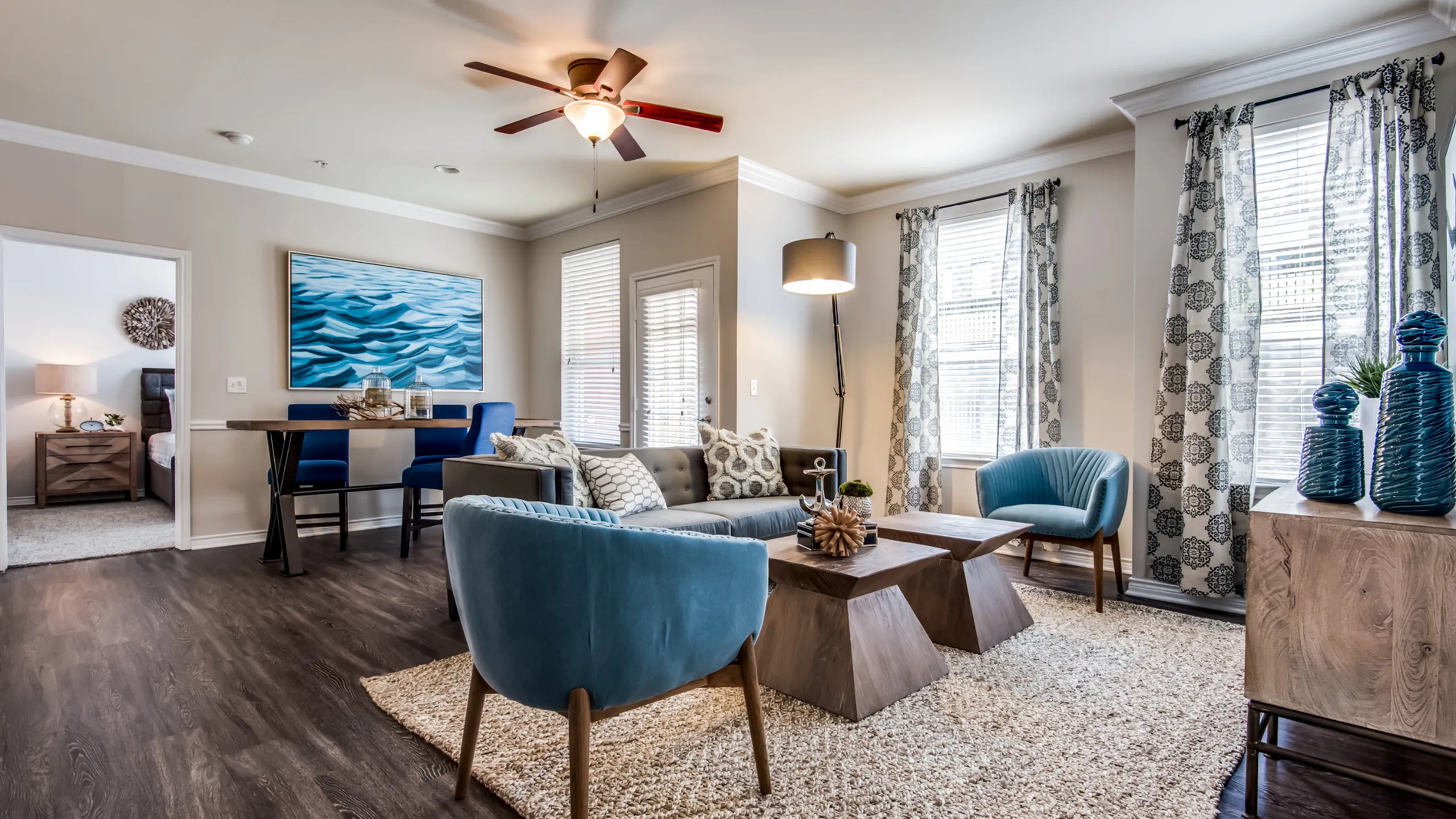 Inviting living space with elegant decor, featuring a cozy gray sofa, chic blue armchairs, and stylish wooden coffee tables, enriched by natural light through large windows and blue accents.
