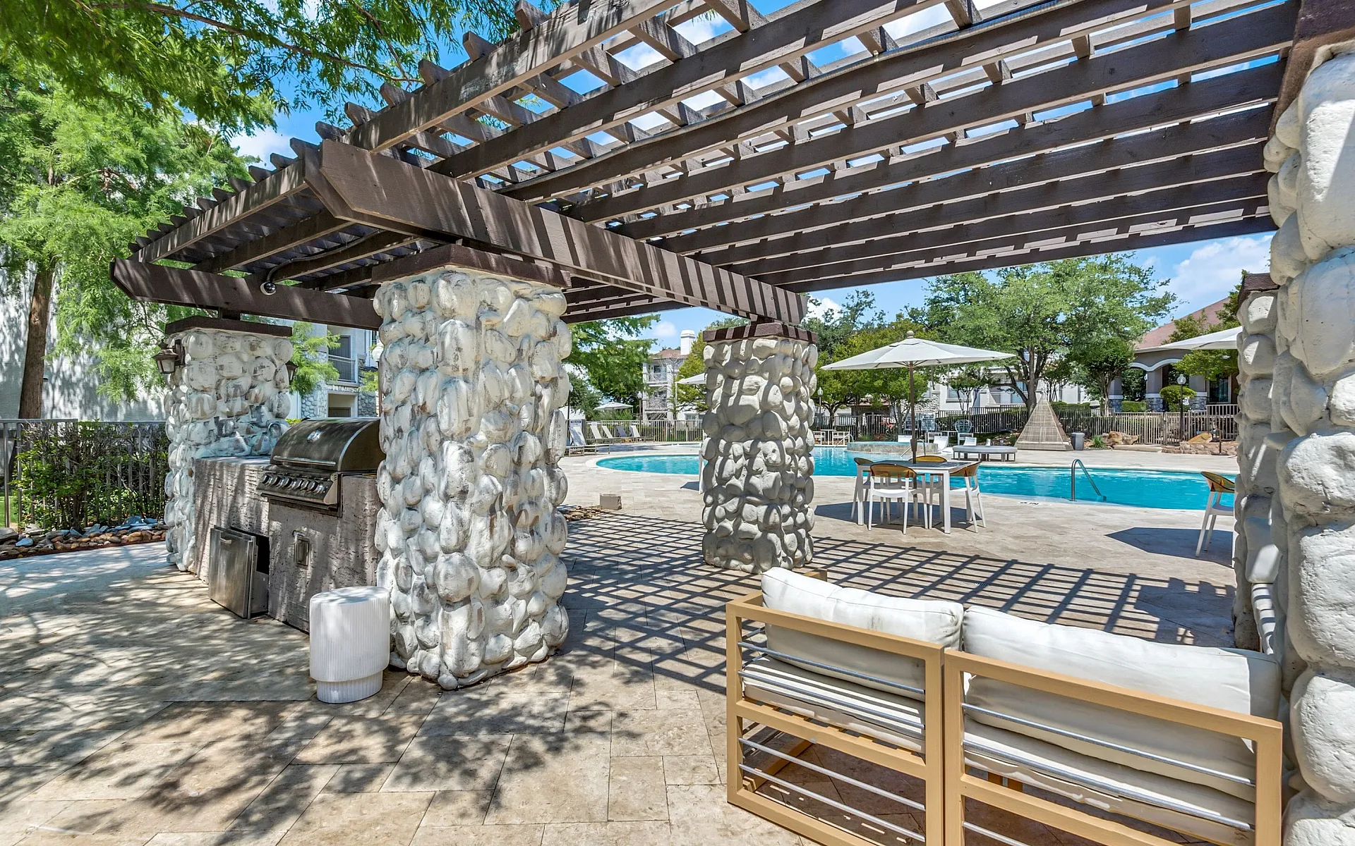 Charming outdoor space featuring a stone-pillared pergola, modern grill, and inviting pool area with sunlit seating.