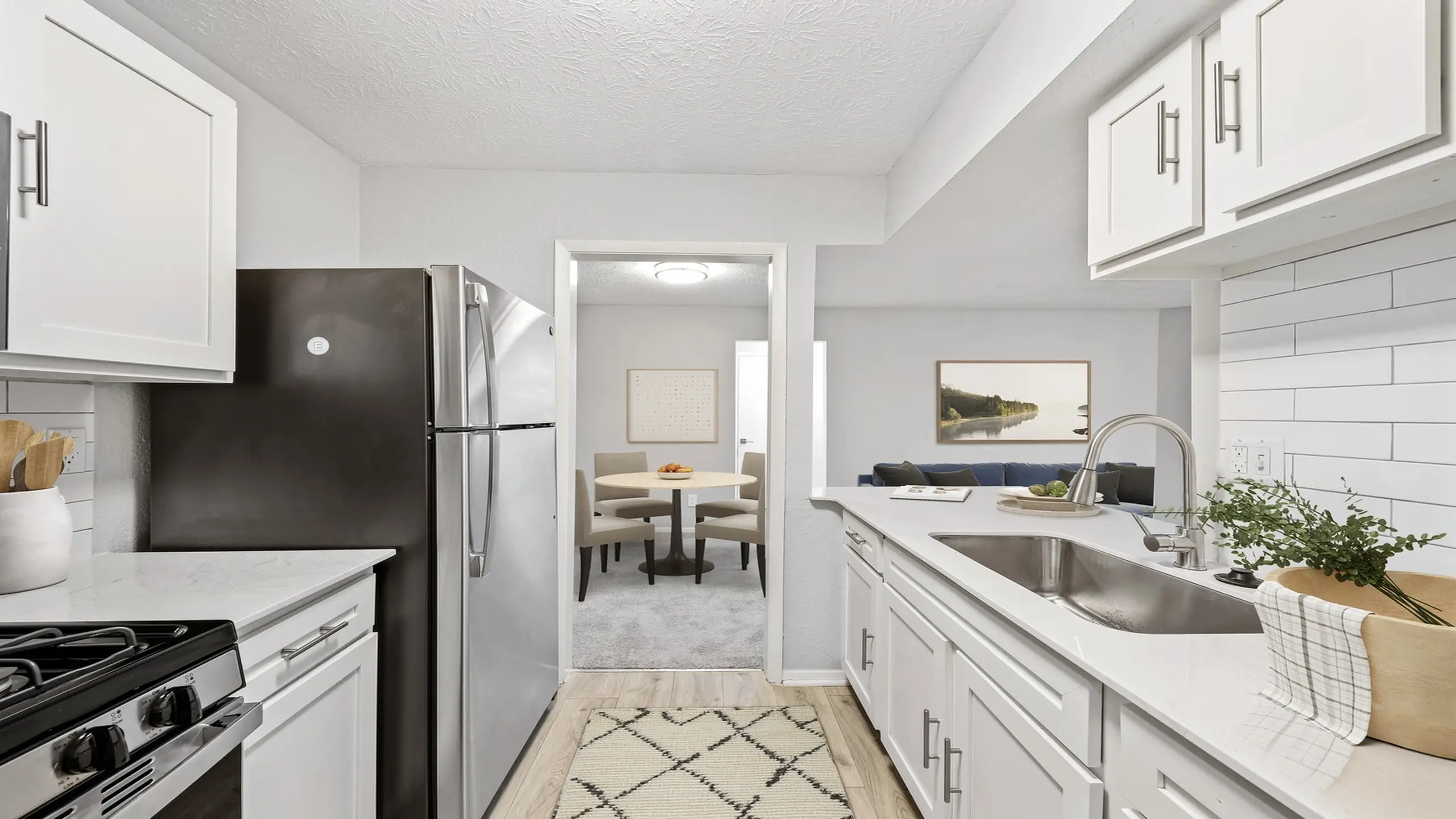 Open-concept apartment kitchen with stainless steel appliances, quartz countertops, white shaker cabinets, subway tile backsplash, and view into dining and living area.
