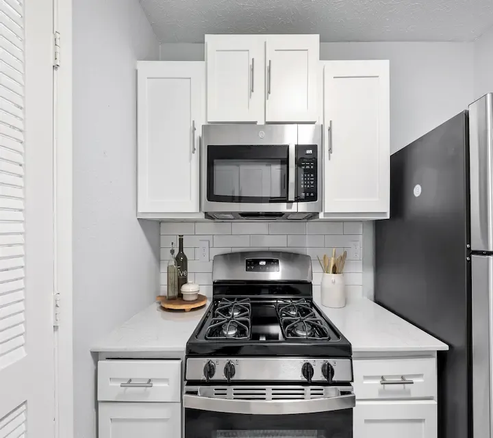 Modern apartment kitchen with stainless steel gas range, built-in microwave, white shaker cabinets, subway tile backsplash, and quartz countertops.