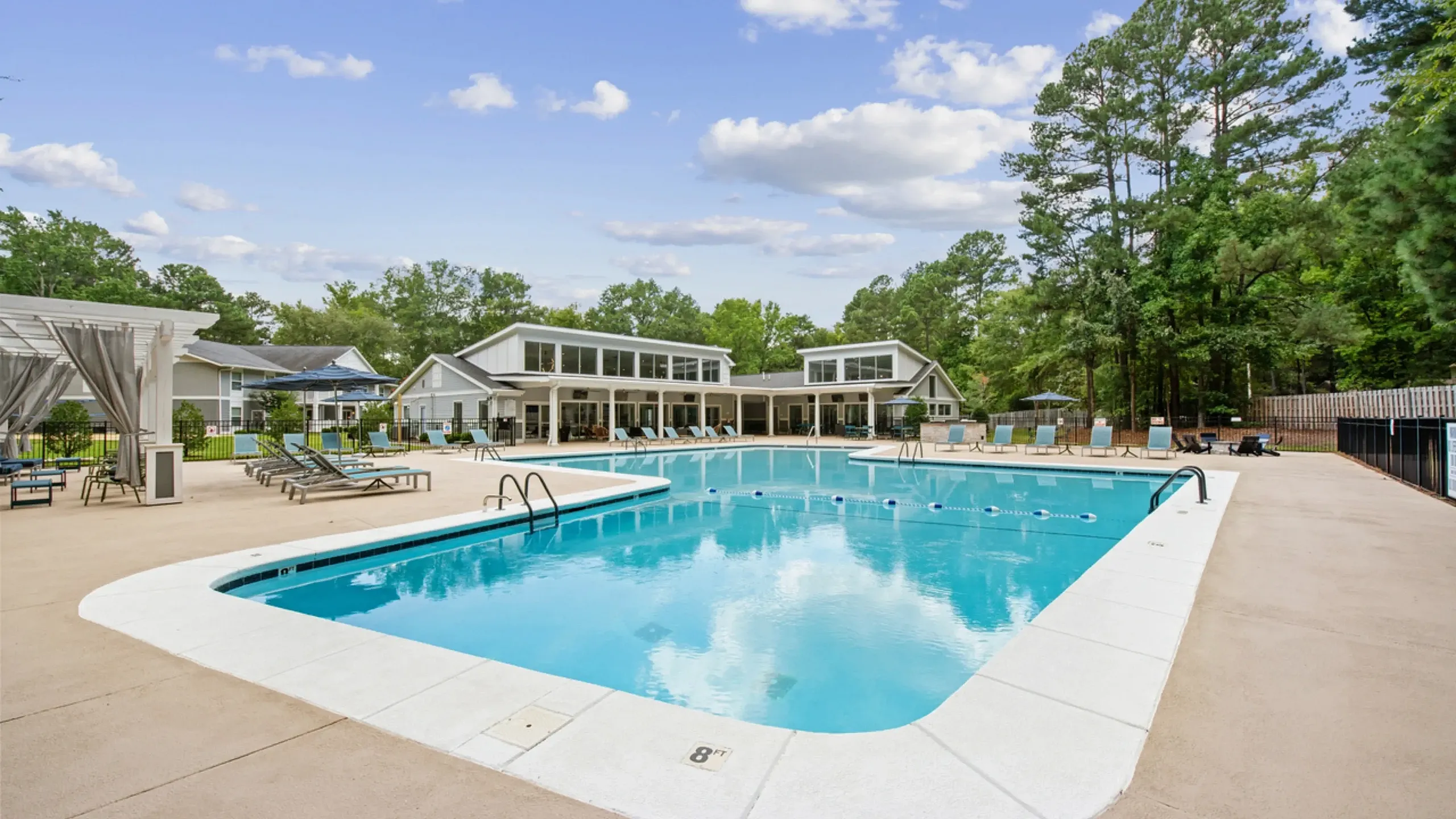 Large outdoor swimming pool with surrounding lounge chairs and umbrellas, set in front of a modern clubhouse and bordered by trees.