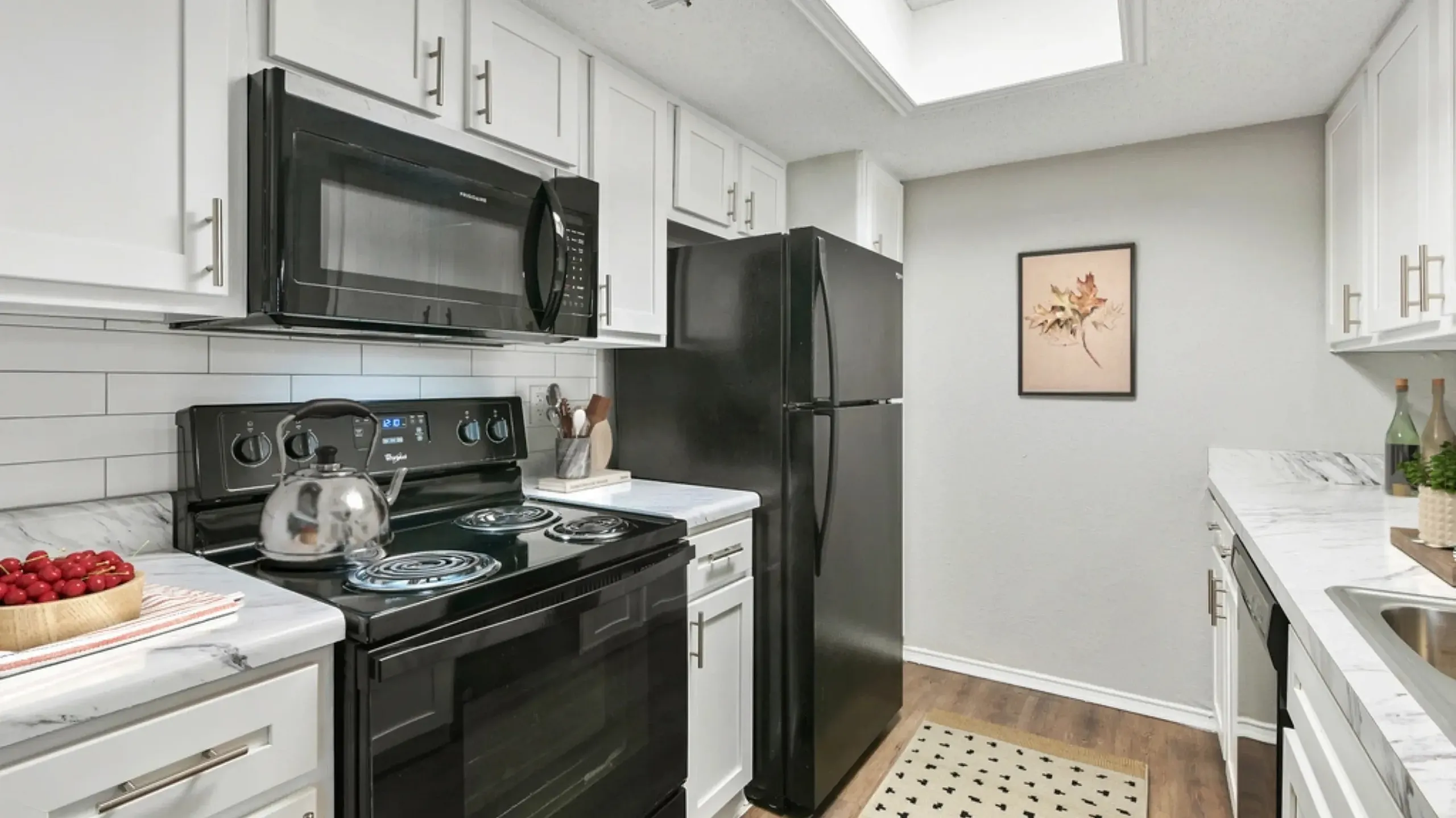 Kitchen with white cabinets, marble-style countertops, and black appliances, including an electric stove, microwave, and refrigerator. Features a subway tile backsplash, wood-style flooring, a small patterned rug, and simple decor with a fr
