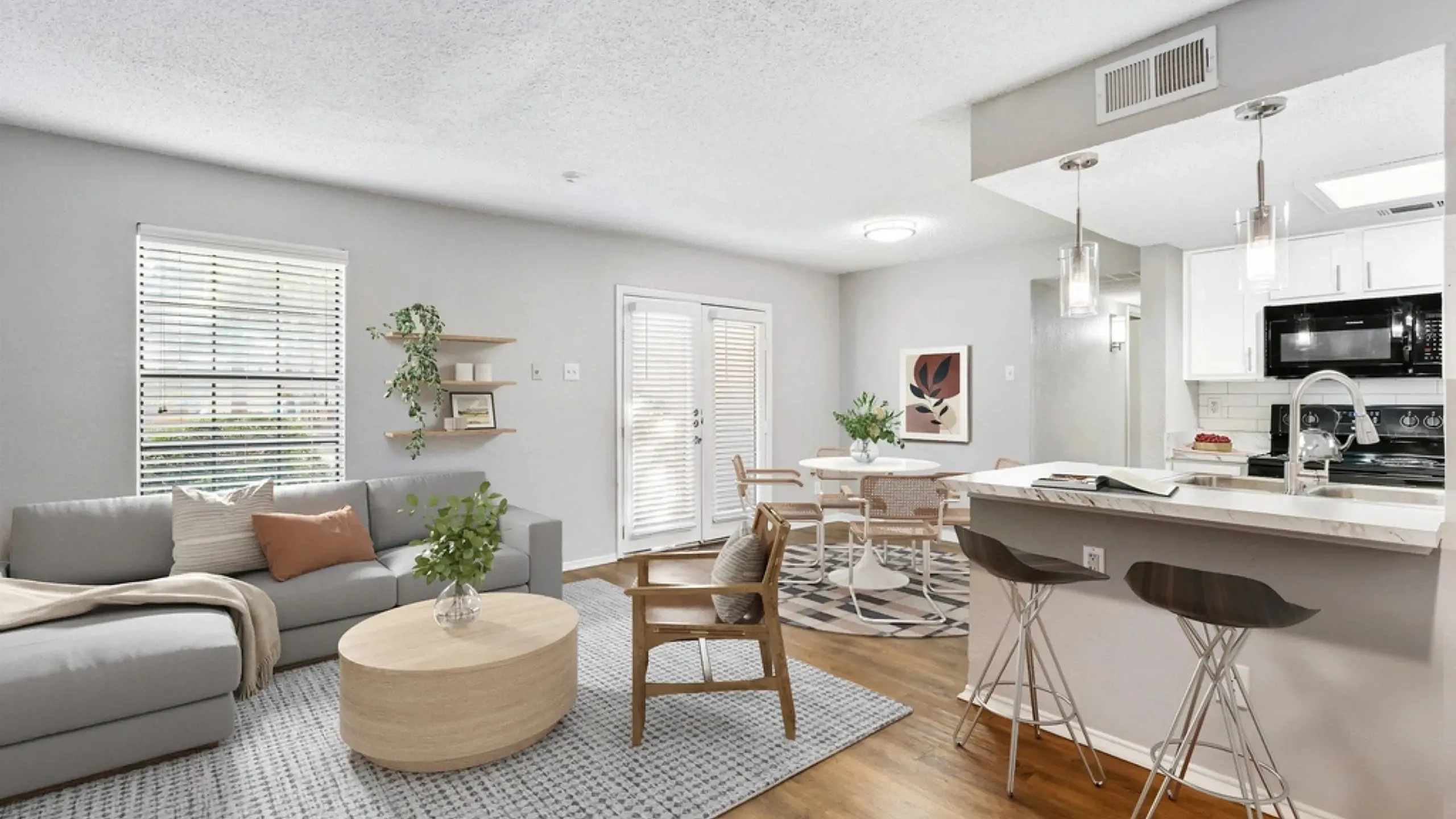 Bright open-concept living and dining area with a gray sofa, round coffee table, and accent chair on a textured rug. The space flows into a dining area with a small table and chairs, and a kitchen with a breakfast bar, pendant lighting, and
