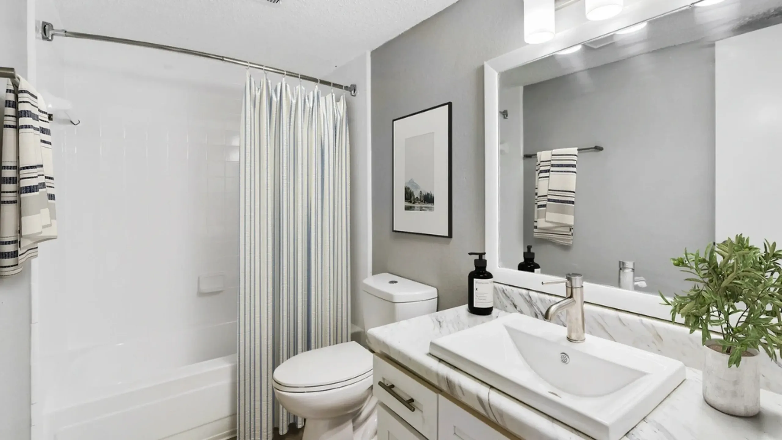 Modern bathroom with a white tub and striped shower curtain, toilet, and a marble-top vanity with a rectangular sink, large mirror, and three-light fixture above. Neutral gray walls, a small plant, and minimal decor create a clean, contempo
