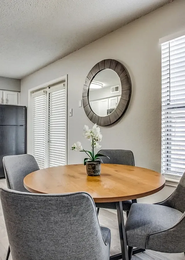 Dining area with hardwood-style flooring, featuring a round wood table and four upholstered chairs. The space connects to a kitchen with white cabinets, marble-style countertops, and black appliances, with large windows, a french door with