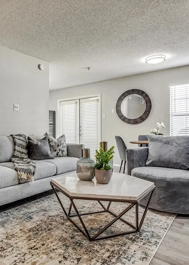 Open-concept living and dining area with hardwood-style flooring, featuring a gray sofa, accent chair, and a coffee table with decor accents on a patterned rug. The space connects to a dining area with a round table and chairs, with large w