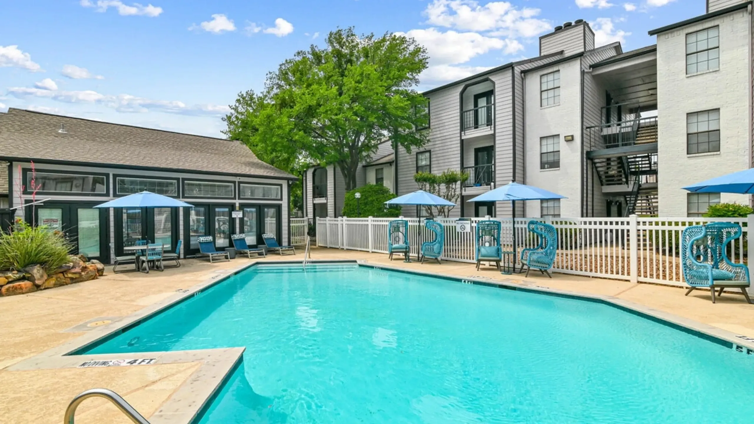 Outdoor community swimming pool with a spacious deck, surrounded by lounge chairs, blue umbrellas, and stylish seating. Features include a white fence, a clubhouse building, and landscaped greenery, with apartment buildings in the backgroun