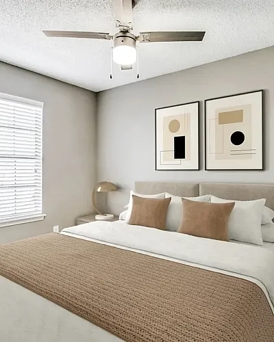 Bright bedroom with plush carpeting, a neatly made bed featuring neutral bedding and accent pillows, and a ceiling fan with light fixture. Features include a window with blinds, matching nightstands with modern lamps, and framed abstract wa