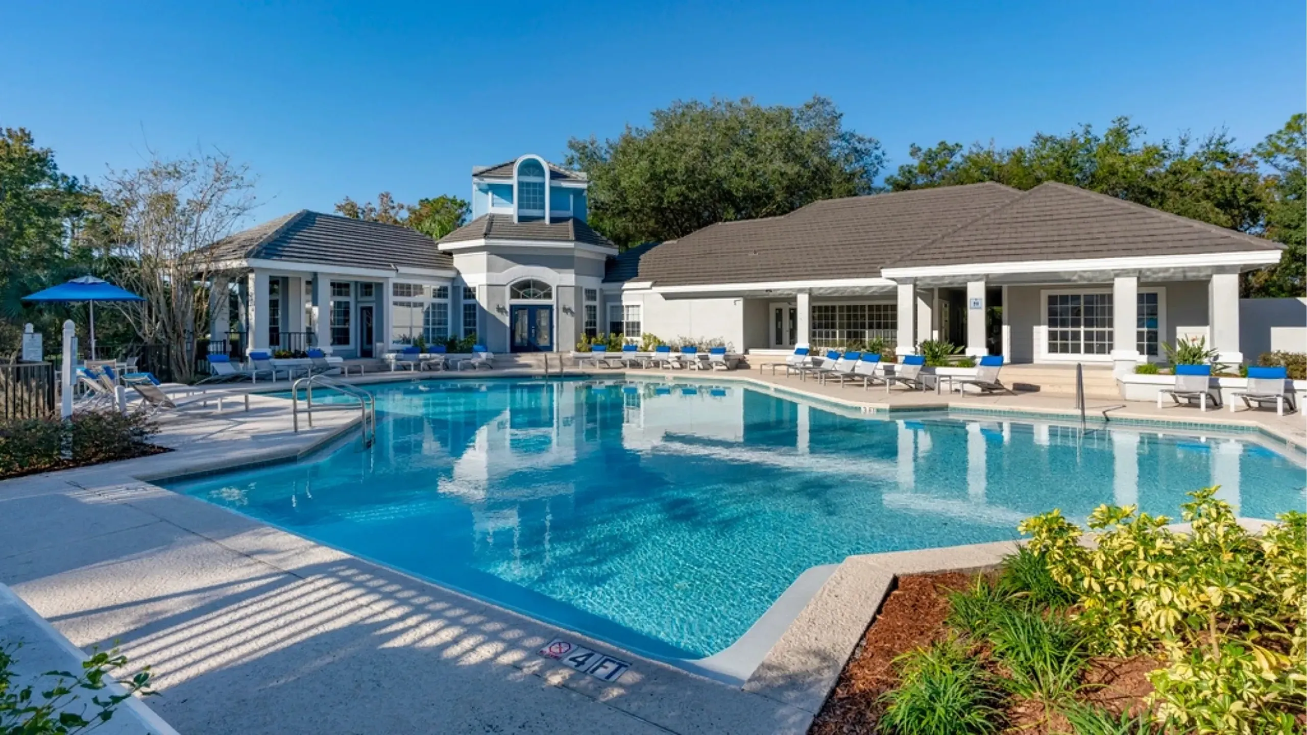 Resort-style swimming pool with surrounding lounge seating and a modern clubhouse, set within landscaped grounds under a clear blue sky.