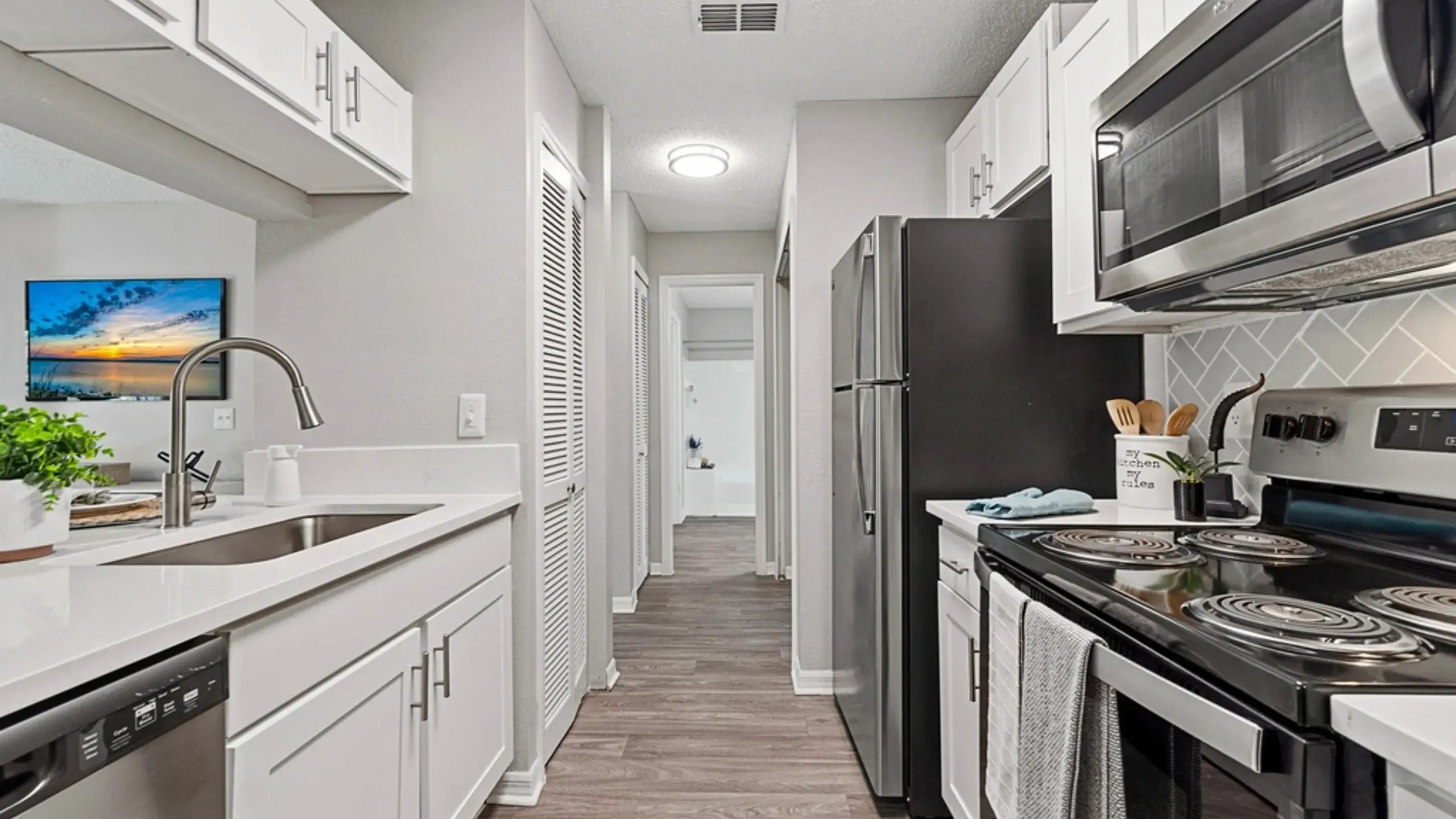 Modern galley-style kitchen with white cabinetry, stainless steel appliances, quartz countertops, and a subway tile backsplash, leading into a hallway beyond.