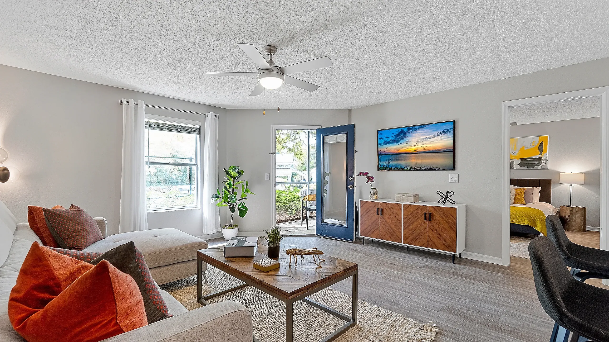 Bright living room with modern furnishings, featuring a ceiling fan, cozy seating, wood-style flooring, and an open door leading to a private patio, with a bedroom visible beyond.