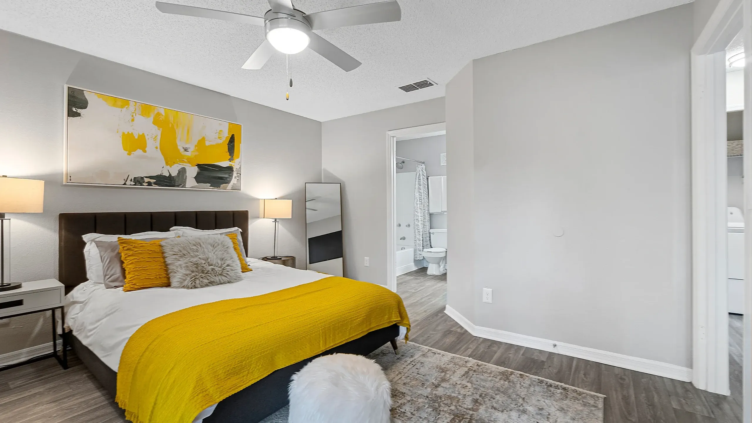 Modern bedroom with a plush bed and yellow accent bedding, featuring a ceiling fan, bedside lamps, and an open layout leading to an en-suite bathroom and closet area.