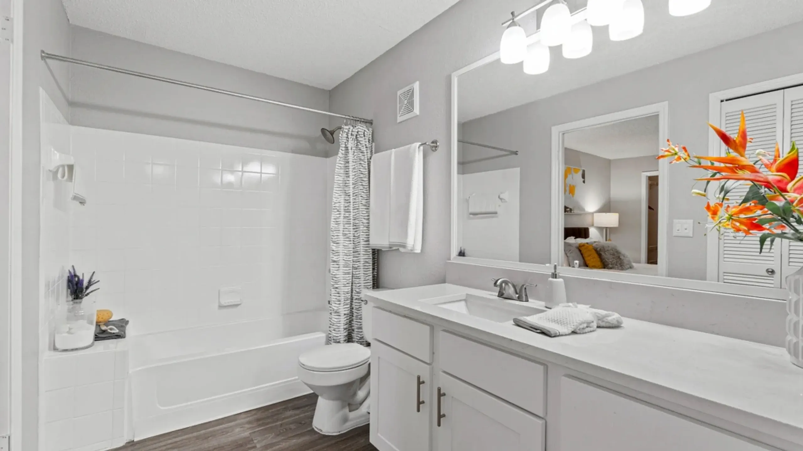 Modern bathroom with a full-size tub and shower, white vanity with ample counter space, large mirror with lighting, and wood-style flooring.