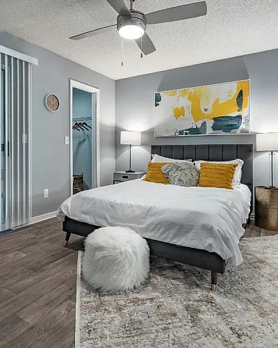 Spacious bedroom with a modern bed and neutral d&eacute;cor, featuring a ceiling fan, sliding glass doors with vertical blinds, bedside lighting, and a floor mirror.