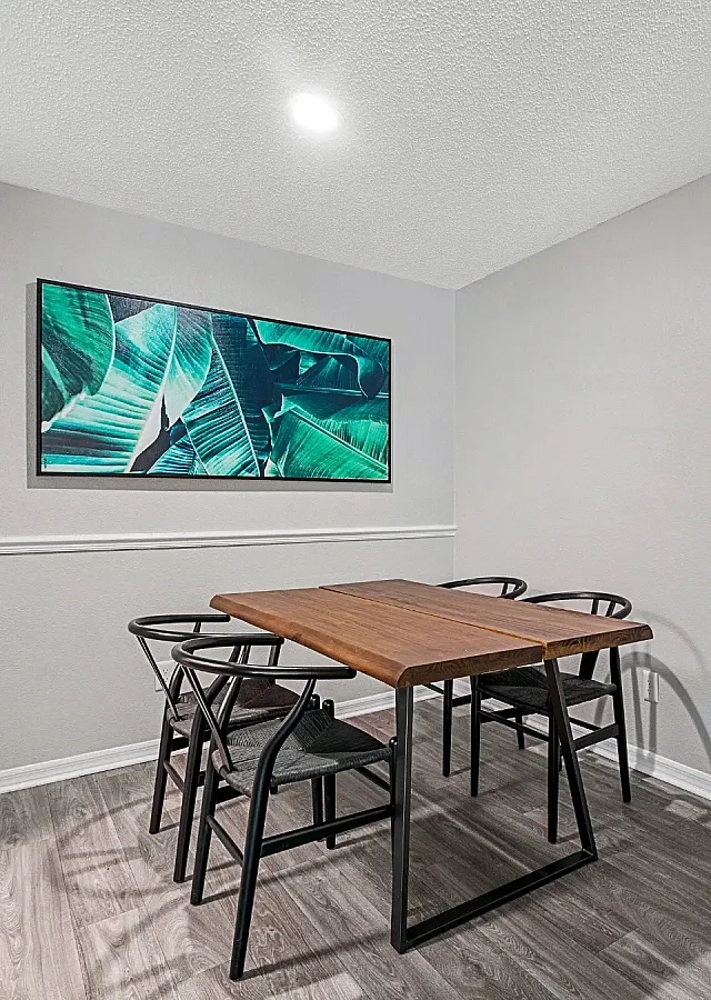 Dining area with a modern table and seating for four, featuring wood-style flooring, neutral walls, and a nearby entry door.