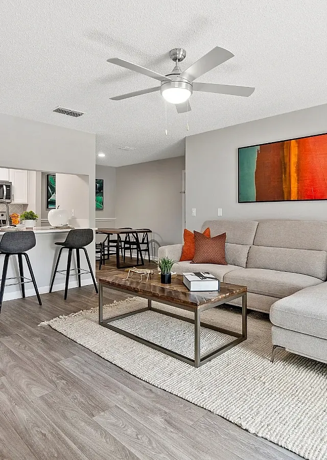 Open-concept living room with a modern sectional sofa, coffee table, and ceiling fan, connected to a breakfast bar and kitchen, with wood-style flooring throughout.