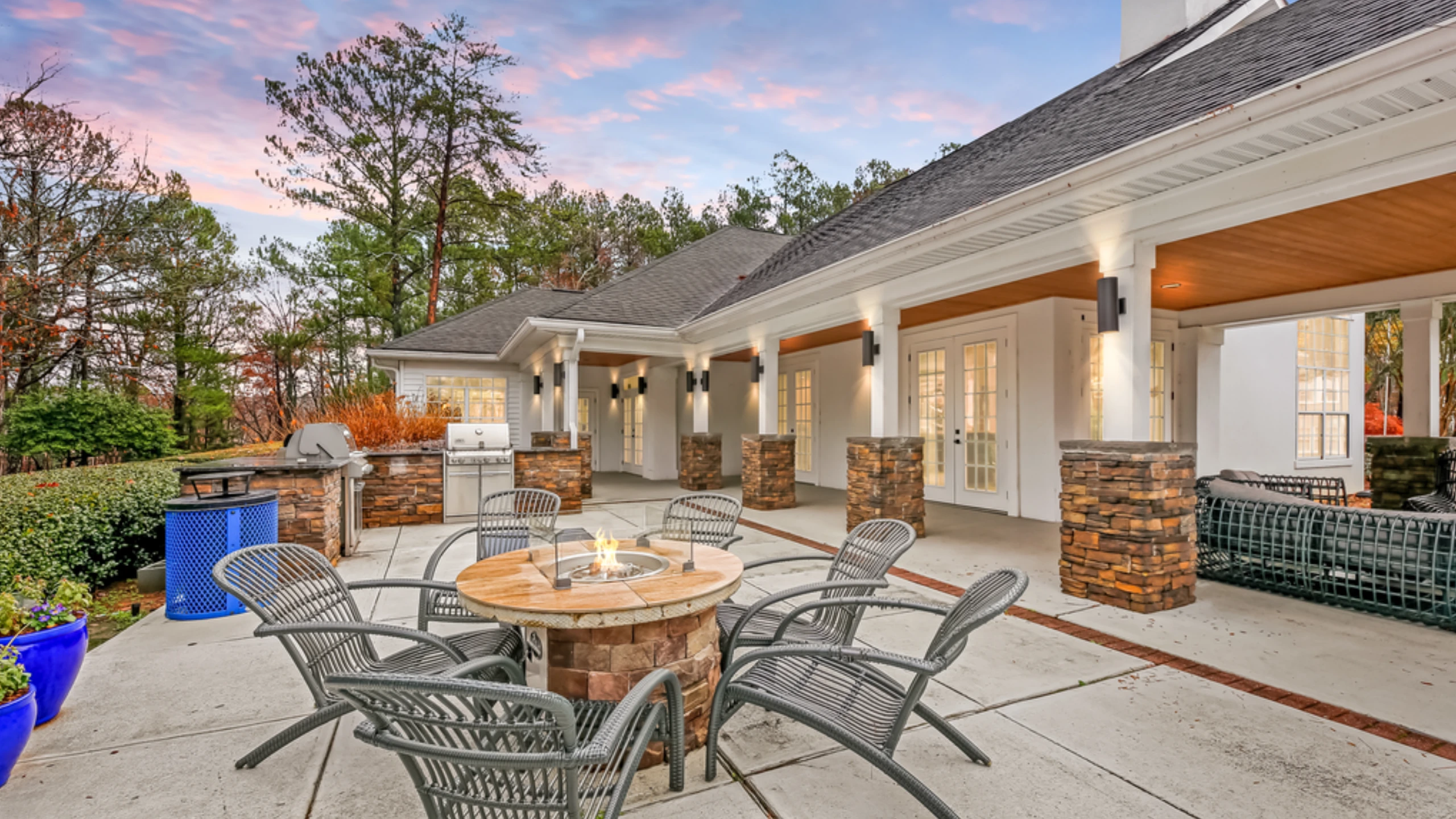 A spacious outdoor resident lounge featuring a circular fire pit surrounded by woven lounge chairs, built-in grilling stations, and a covered clubhouse patio with stone-accent columns and warm lighting. The space overlooks landscaped greene
