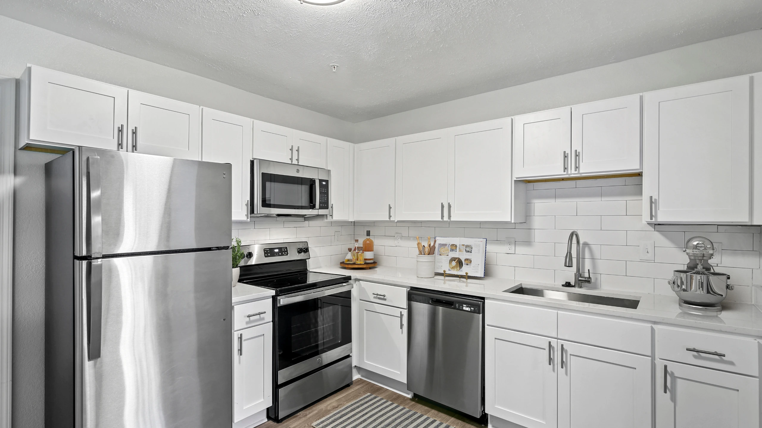 Modern apartment kitchen with white shaker-style cabinets, white subway tile backsplash, quartz-style countertops, and stainless steel appliances including a refrigerator, microwave, oven, and dishwasher. The space also features a deep unde
