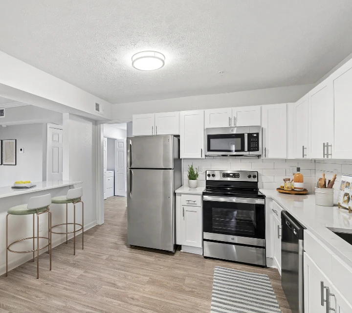 Bright modern kitchen with white shaker-style cabinets, stainless steel appliances, white subway tile backsplash, and wood-style flooring. The space features a breakfast bar with seating, quartz-style countertops, contemporary finishes, and