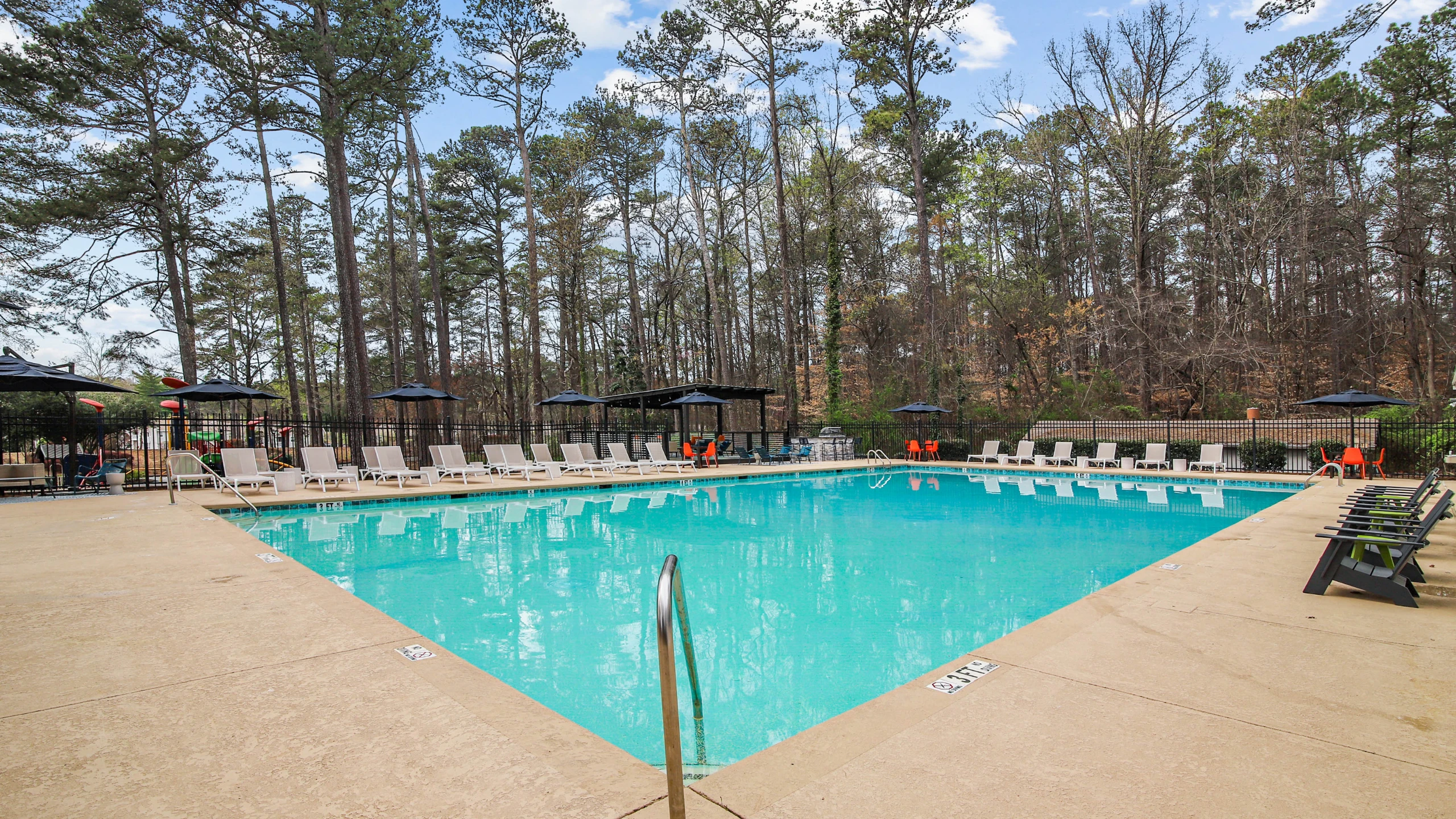 Large outdoor swimming pool surrounded by lounge chairs, umbrellas, and tall pine trees in a peaceful wooded setting. The resort-style pool area features a spacious concrete deck, shaded seating areas, and a relaxing atmosphere under a brig