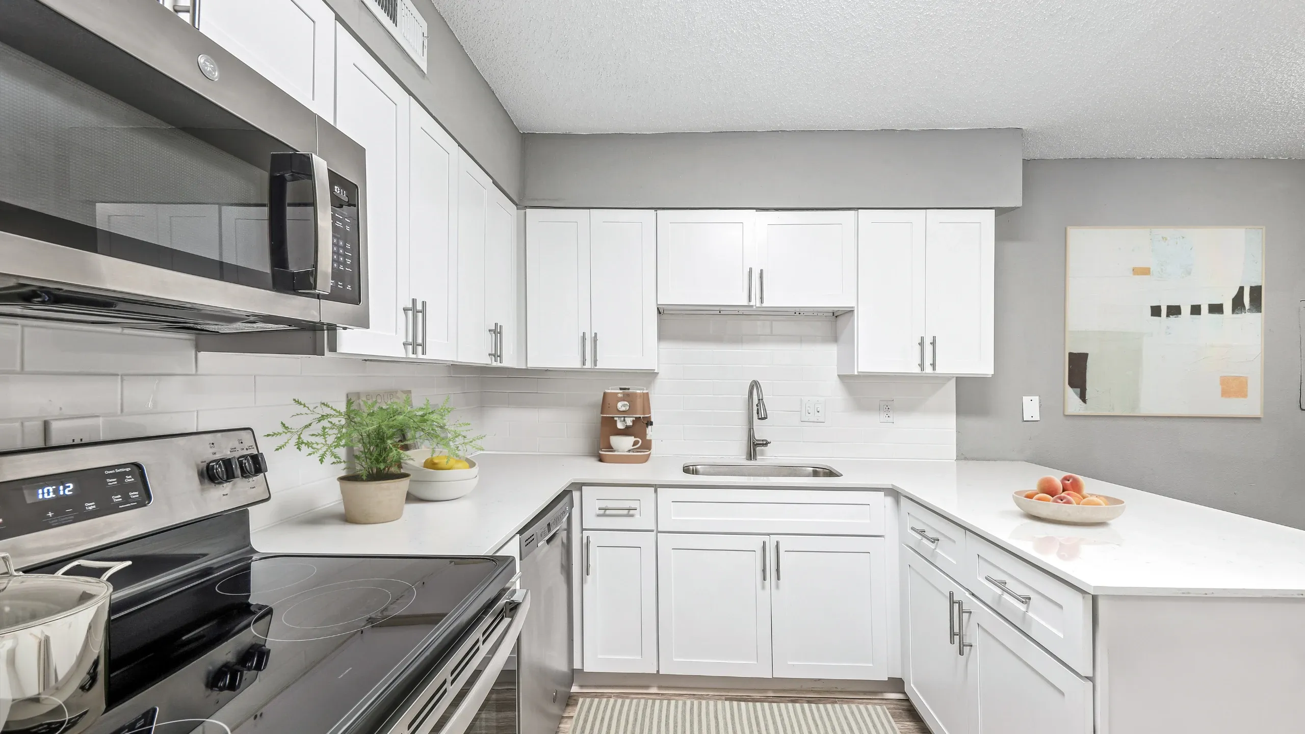 Bright modern kitchen with white shaker-style cabinets, white countertops, and stainless steel appliances. The space features a built-in microwave, electric stove, dishwasher, and under-mount sink with a sleek faucet. Light gray walls, subw