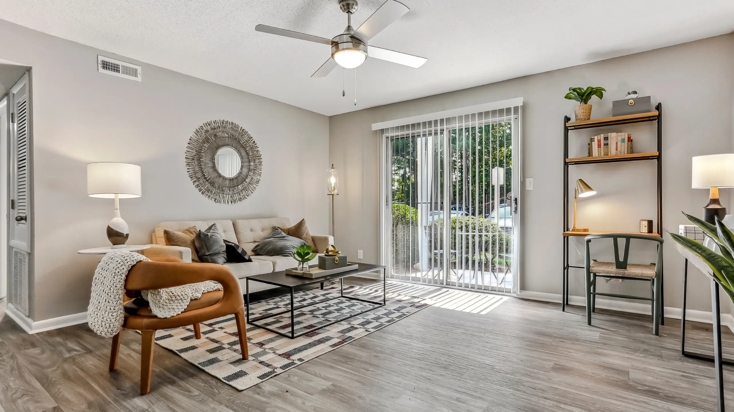 Modern living room with neutral-toned decor, featuring a beige sofa, patterned area rug, wood-look flooring, and a cozy accent chair. Large sliding glass doors allow natural light into the space and lead to a private patio area. A small des