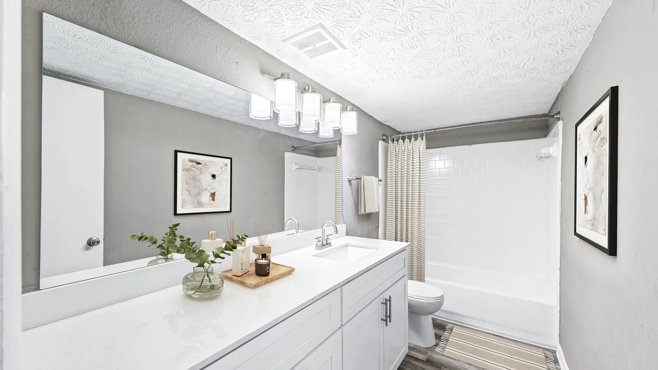 Modern bathroom with a long white vanity, sleek countertop, and large wall mirror with contemporary light fixtures. The space features gray walls, white cabinetry, a bathtub and shower combination with a striped curtain, and minimalist deco